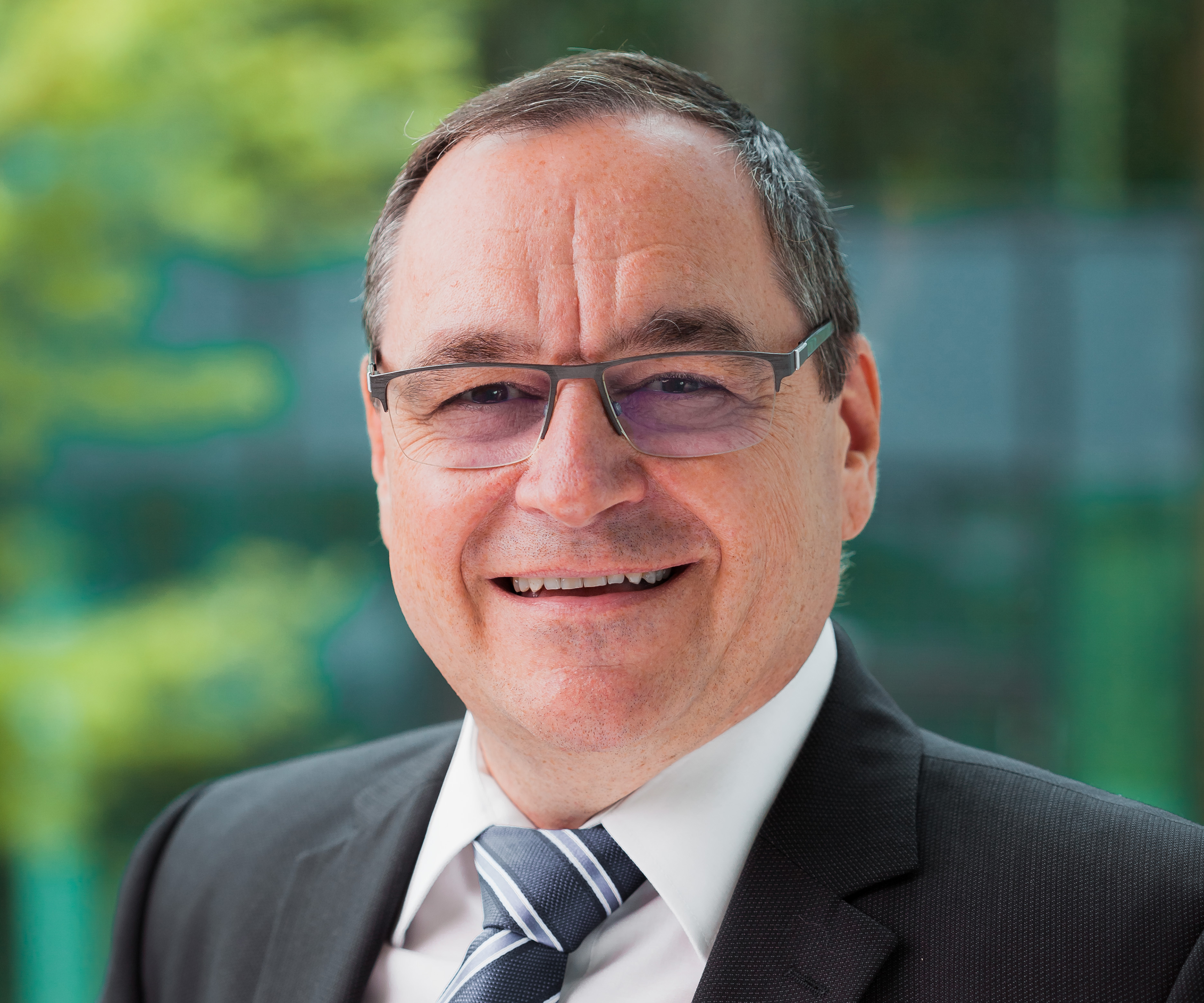 Ein lächelnder Mann mit Brille und kurzen Haaren trägt einen dunklen Anzug, ein weißes Hemd und eine gestreifte Krawatte. Der Hintergrund ist mit Grün verschwommen, was eine professionelle Umgebung im Freien suggeriert.