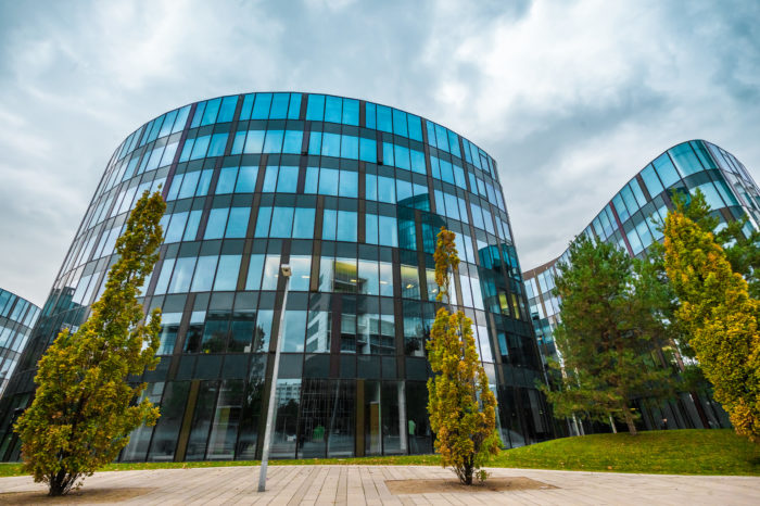 Ein modernes Bürogebäude aus gebogenem Glas mit reflektierender Fassade unter einem bewölkten Himmel. Das Gebäude ist von einigen hohen, belaubten Bäumen umgeben und im Vordergrund befindet sich ein gepflasterter Gehweg.