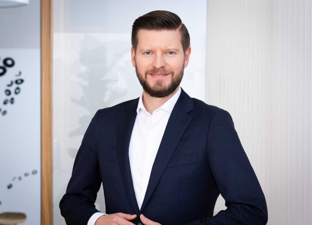 Mann mit kurzen braunen Haaren und Bart, trägt ein weißes Hemd und ein dunkles Jackett und lächelt in die Kamera. Der Hintergrund ist sanft verschwommen und weist helle Grautöne auf.