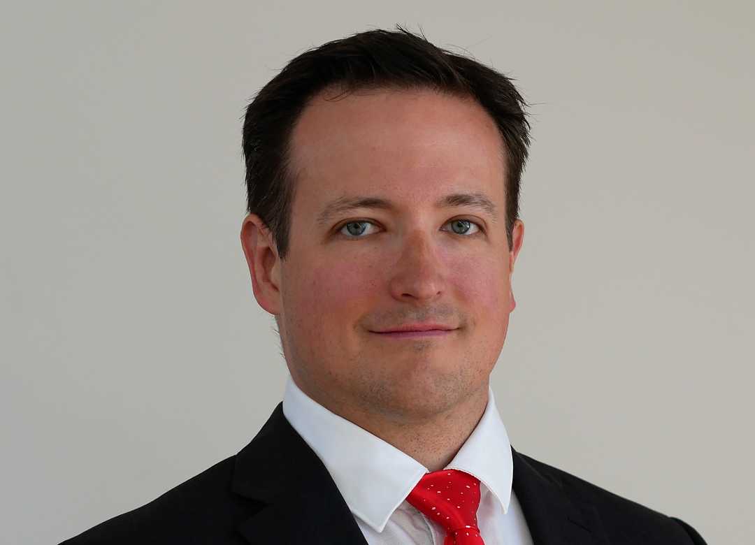 Ein Mann in Anzug und roter Krawatte steht vor einem schlichten Hintergrund und blickt mit neutralem Gesichtsausdruck direkt in die Kamera. Er hat kurzes dunkles Haar und ein glattrasiertes Gesicht.